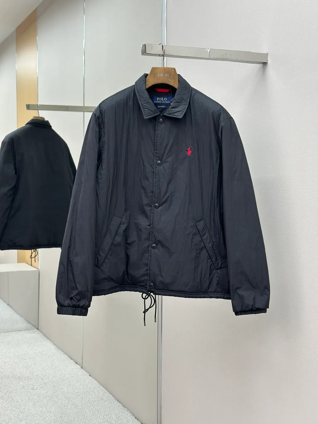 P650 RAH LAUREN RL red label lapel is light and thin. It is made of high-density nylon taffeta with a density of about 210T-300T. The surface is smooth and slightly matte. It has excellent windproof and waterproof performance (with R water-repellent treatment). It has strong wrinkle resistance and is easy to care for. It adopts the vat dyeing process (the dye penetrates into the fiber) and the color fastness reaches ISO level 4 or above to avoid fading or color transfer after washing. The lightweight cotton-polyester blend lining (65% Polyester + 35% Cotton) reduces the friction between the fabric and inner clothing and improves the smoothness of wearing. The blended material combines breathability and durability to avoid the stuffy feeling of pure nylon lining. The classic coach (Jaet) loose fit has a slightly dropped shoulder line (the shoulder width is 1-2cm wider than the standard fit), the length reaches the hips, and the sleeves are slightly tapered straight sleeves, taking into account freedom of movement and casual style. Flat satin stitch embroidery (satin embroidery), using 100% polyester high color fastness embroidery thread (the color number is consistent with the brand's standard red color card), the stitch density is 12-15 stitches cm, the outline of the horse logo is clear, and the details of the rider and horse are burr-free. Precise positioning through the template (12cm from the bottom of the collar and 8cm from the placket) ensures symmetry and centering. The seams of the body are made of double-needle flat seams (needle pitch 3cm, 10 needles), the stitches are flat and straight, the stitching color is dark black (the same color as the fabric), and the stitch width is 0.5cm, which enhances firmness and forms simple decorative lines. The placket is made of double-layer folding + flat seam splicing, with a thickness of about 0.3cm to ensure that the buttons are not easily deformed when fixed; the edges of the placket are cut with a hot knife to avoid burrs. The armholes are machine stitched with round head keyholes and flat seams. After stitching, the armholes have a natural curvature and no sense of tightness during movement. The height of the sleeves is about 8-10cm, which is in line with the casual and loose characteristics. Black urea resin button with matte surface treatment. The buttonholes are hand-made keyholes (6 strands of cotton thread, keyhole density of 8 stitches mm), and the edges of the eyelets are smooth and without burrs. The first button on the neckline is a hidden button (diameter 12mm), which prevents the neckline from opening and improves the neatness of wearing. Size:S-XL